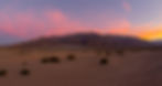 Death Valley Mesquite Dunes desert landscape at sunset, with a mountain range in the background. Sky is pink and orange, creating a serene, warm mood. Sparse bushes dot the sand.