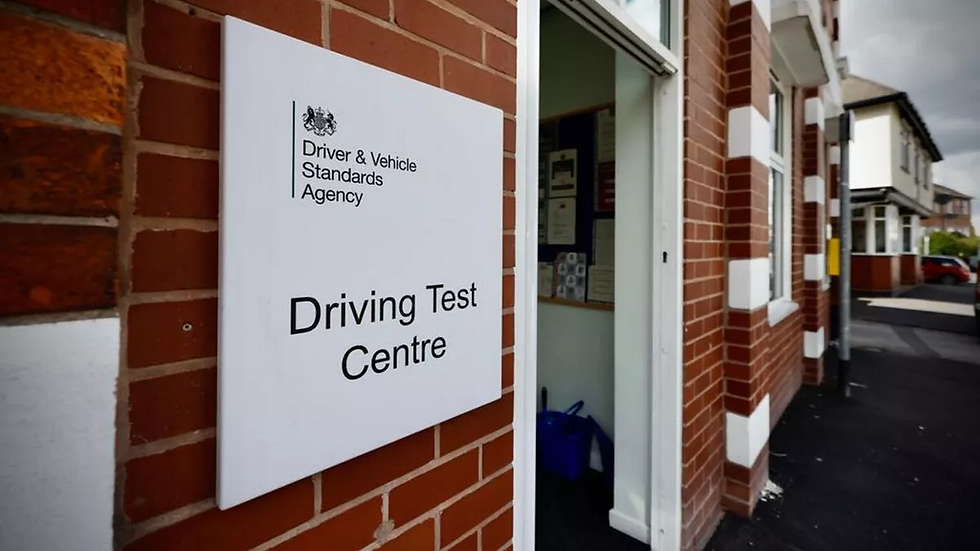 Sign for a Driving Test Centre on a brick wall. Open door leads inside. Overcast sky. Building has red bricks with white accents.