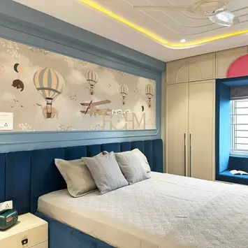 Son’s bedroom interior designed by hohm editors, interior designers in Hyderabad, featuring a bold blue accent wall with playful hot air balloon motifs, platform bed with layered white bedding and grey pillows, integrated bedside table, built-in wardrobe with soft beige and red panels, large window with blue frame, recessed ceiling with yellow cove lighting, and modern ambient decor—creating a vibrant, imaginative, and comfortable personal retreat for a child or teenager.