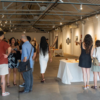 O espaço expositivo está cheio de visitantes. Pessoas circulam entre as obras e mesas, algumas em grupos conversando animadamente. As paredes brancas exibem quadros e instalações, e o teto metálico com iluminação direcionada realça o clima de vernissage.