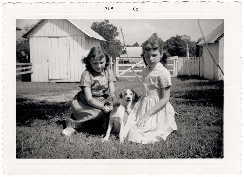 Sisters Beth (now Fiddler) and Mary Ruth Yoe with puppies; farm outbuildings in background, 1960.