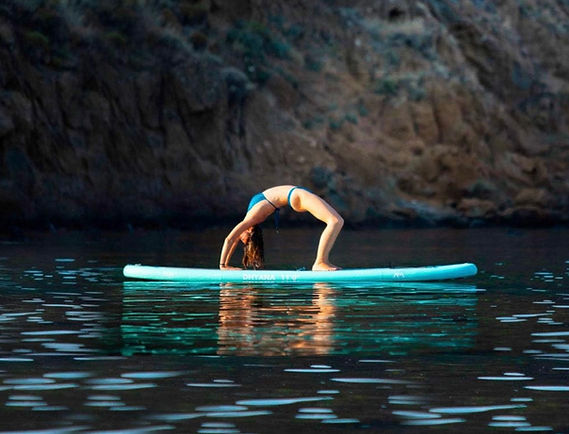 A woman in a bikini does a backbend yoga pose on a light blue paddleboard in calm water