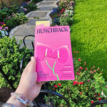 A hand holds up a paperback copy of hunchback by Saou Ichikawa. The background behind the book shows a green garden in the midst of summer with a stone path. The book cover is bright pink with lillies on the cover also in pink