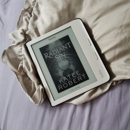 A kobo e-reader rests on top a cream frilly cushion cover and a worn out purple matress cover.