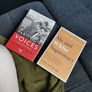 Two paperbacks rest on the seat of a grey sofa. There is a green coloured cardigan in the corner of the image. One of the paperback books is red on the bottom half of the cover and has an image of men standing by a metal fence. In white letters the title reads Partition Voices. The second book is pale orange in colour with white and black bold text reading Me and White Supremacy.