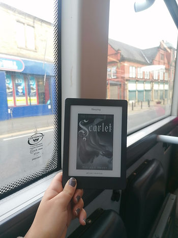 A hand holds up a kobo aura e-reader. In the background there is the window of a bus,a road, and a betting shop. The face of the e-reader shows the book cover for Scarlet by Marissa Meyer.