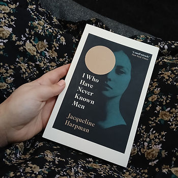 A hand holds a paperback book of I Who have Never Known Men by Jacqueline Harpman. The cover is dark with a blue tone. An image displays a solemn faced women with a large golden cicrle covering the upper left of her face like a sun. The background shows a the legs of a woman covers in the flower patterned dress of blue, black, gold, and white.