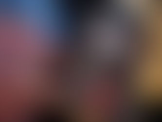 A hand holds a paperback copy of The Hunger Games by Suzanne Collins. The book has a black cover, decorated with bold white lettering and splashes of blood. In the centre is an HG logo with a bird and an arrow. Behind the book is a grey sofa, the lower body of a person wearing a pink jumper and faded blue jeans. 