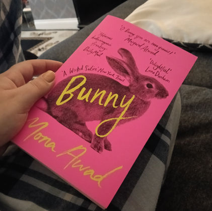 A hand holds a paperback copy of Bunny by Mona Awad. The book is vivid pink in colour and features a wild rabbit on the cover with yellow coloured swirly text. The background shows tartan patterned trouser legs of  a person and a dark grey sofa. 
