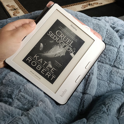 A hand holds a kobo ereader with a pink folding case open to reveal the book cover for the title book Cruel Seduction by Katee Robert. The background of the image shows the ereader is resting on a grey-blue coloured blanket with a cross-hatch pattern.