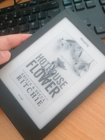 A hand holds an ereader over a brown desk and keyboard. The e-reader shows the book cover for Hothouse Flower by Becca and Krista Ritchie