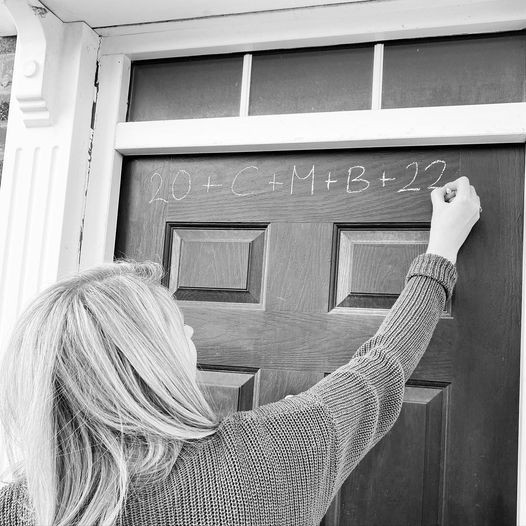 Woman writes blessing in chalk on a door to a home.