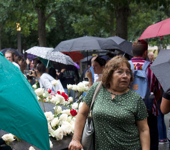 A woman has her hand on the corner of the 9/11 memorial pool as she gazes off frame in what looks like a moment of contemplation. She's framed by onlookers umbrellas and a field of commemorative flowers adorning the memorial space.