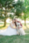 couple de mariés assis dans l'herbe avec leur chien beige. La mariée est en robe blanche et le marié en costume rouge.