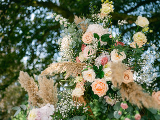 Grand bouquet de roses crème et pêche avec herbes de pampa, en extérieur sous des arbres flous, ambiance romantique.