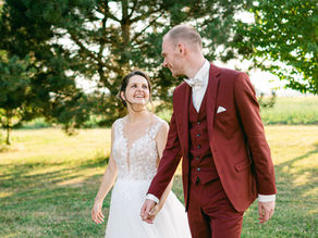 Jeune couple de mariés se tenant la main et souriant dans un parc, mariée en robe blanche, marié en costume bordeaux.