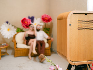 Cabine photo en bois au premier plan, photobooth, couple flou assis sur canapé en rotin entouré de grandes fleurs, ambiance romantique.