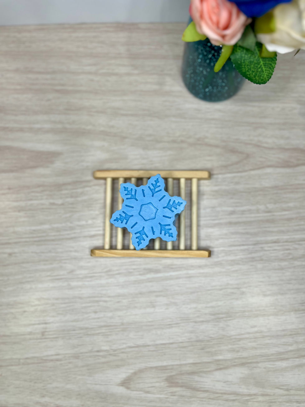 Jaxnrose Soaps Bamboo Soap Dish with a blue snowflake bar of soap placed on it. There is a vase filled with roses and jacks in the background. 