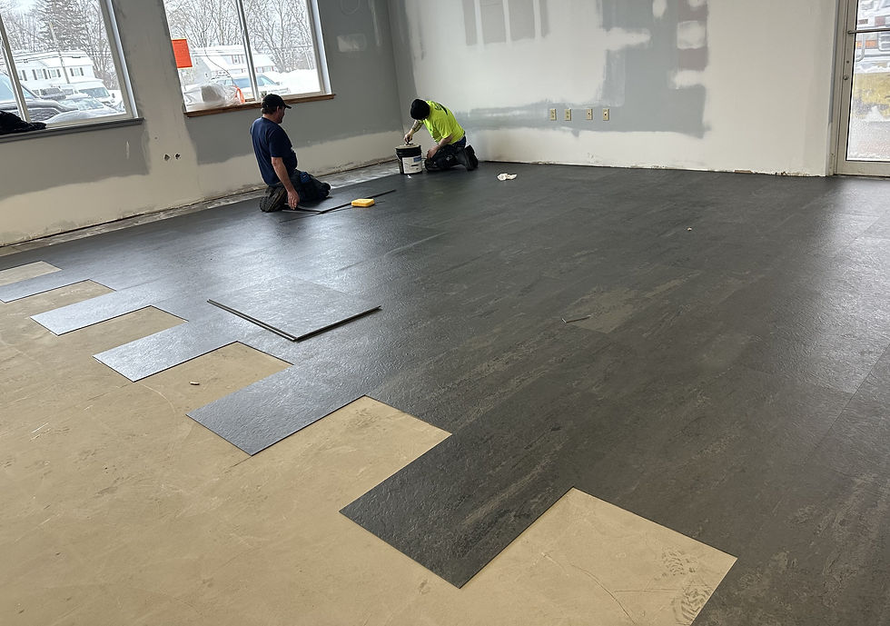 Two workers lay dark vinyl flooring in a bright room with large windows. One applies adhesive from a bucket, while the other kneels nearby.