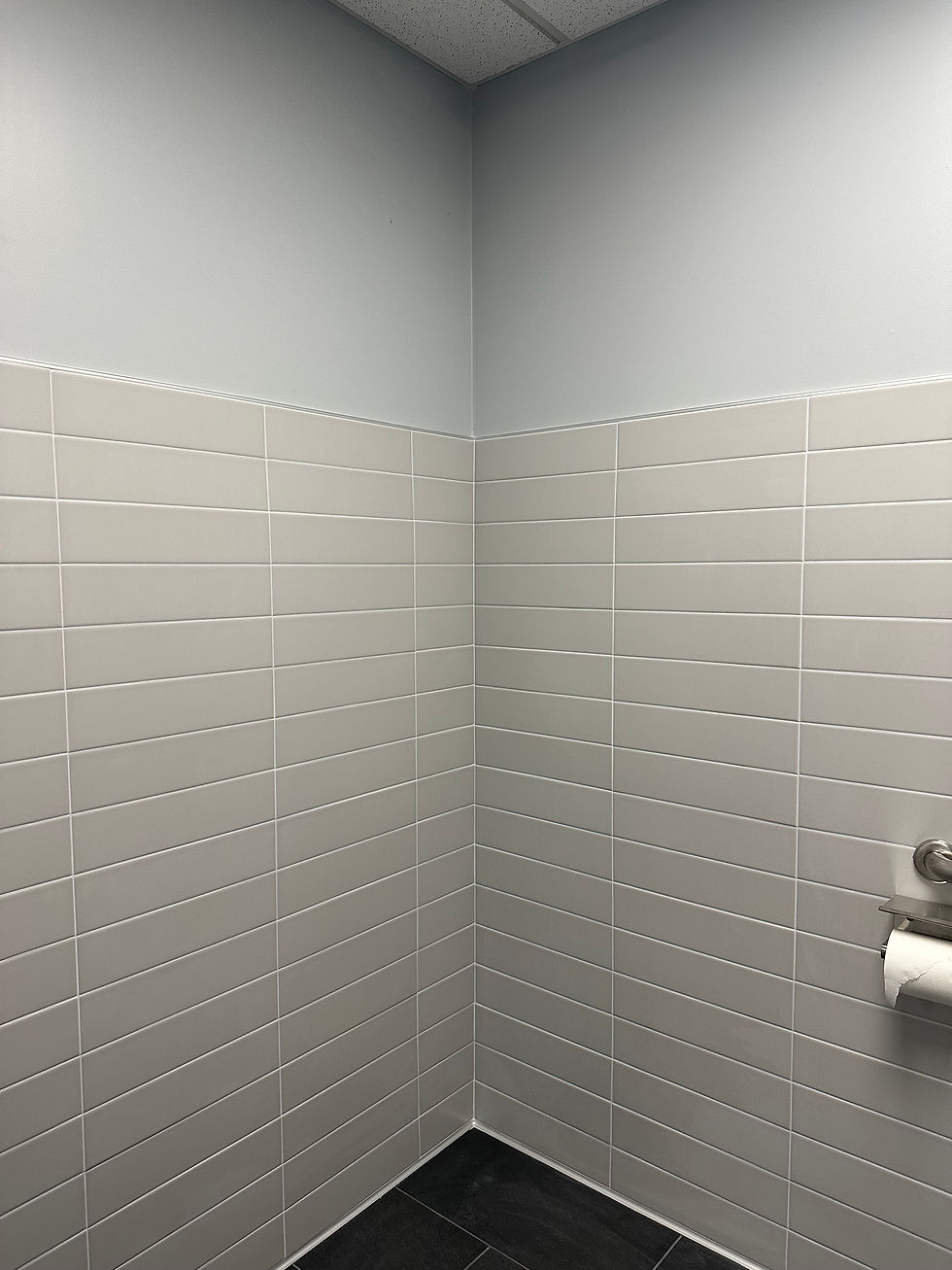 Bathroom corner with light gray tiles, white wall above, and a toilet paper roll on the right. Floor is dark, creating a neutral mood.