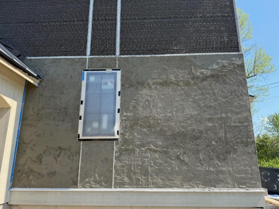 Gray hard stucco house wall under construction with a covered window, black upper siding, and blue sky in the background.
