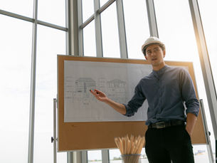 Architect in a hard hat points at house blueprints on a board in a sunlit room with large windows, conveying focus and professionalism.