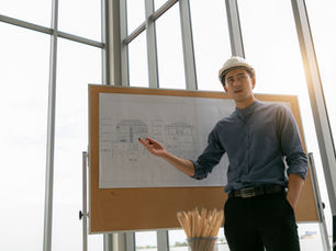 Architect in a hard hat points at house blueprints on a board in a sunlit room with large windows, conveying focus and professionalism.