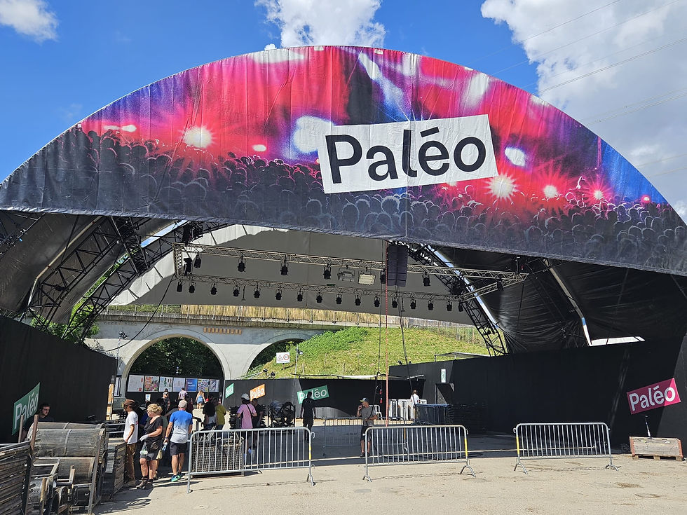 Outdoor music stage setup at the Paleo Festival with people preparing area. Colorful canopy and festival signs. Clear blue sky. Energetic mood.