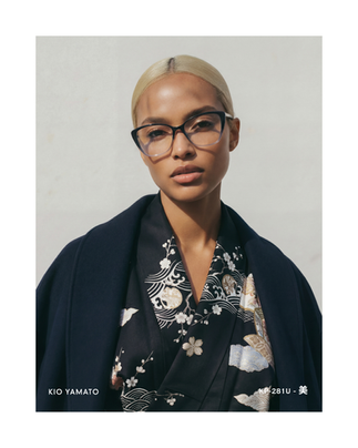 Model wearing KP-281U eyewear by KIO YAMATO, styled in a dark robe with subtle pink accents, photographed front-facing in soft architectural light.