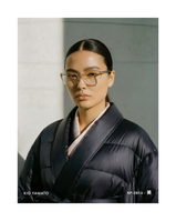Model wearing KP-281U eyewear by KIO YAMATO, styled in a dark robe with subtle pink accents, photographed front-facing in soft architectural light.