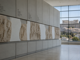 Parthenon frieze fragments and empty sections displayed in the Acropolis Museum, with the Acropolis visible through large windows.