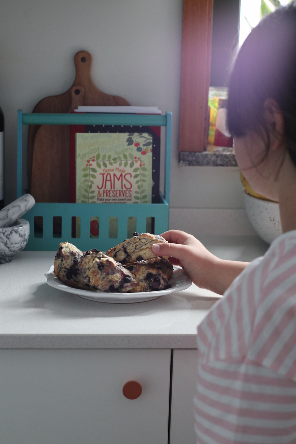 Menina com camiseta listrada pega bolinho em prato sobre bancada. Ao fundo, livro de receitas "Home Made Jams & Preserves". Emocional calmo.