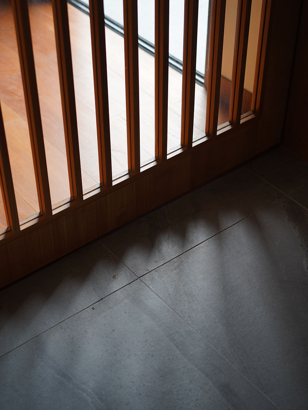 Close-up architectural detail of a custom wooden high door featuring a precise slatted design, showing natural light casting dramatic shadows on the floor of a moody transitional interior space.