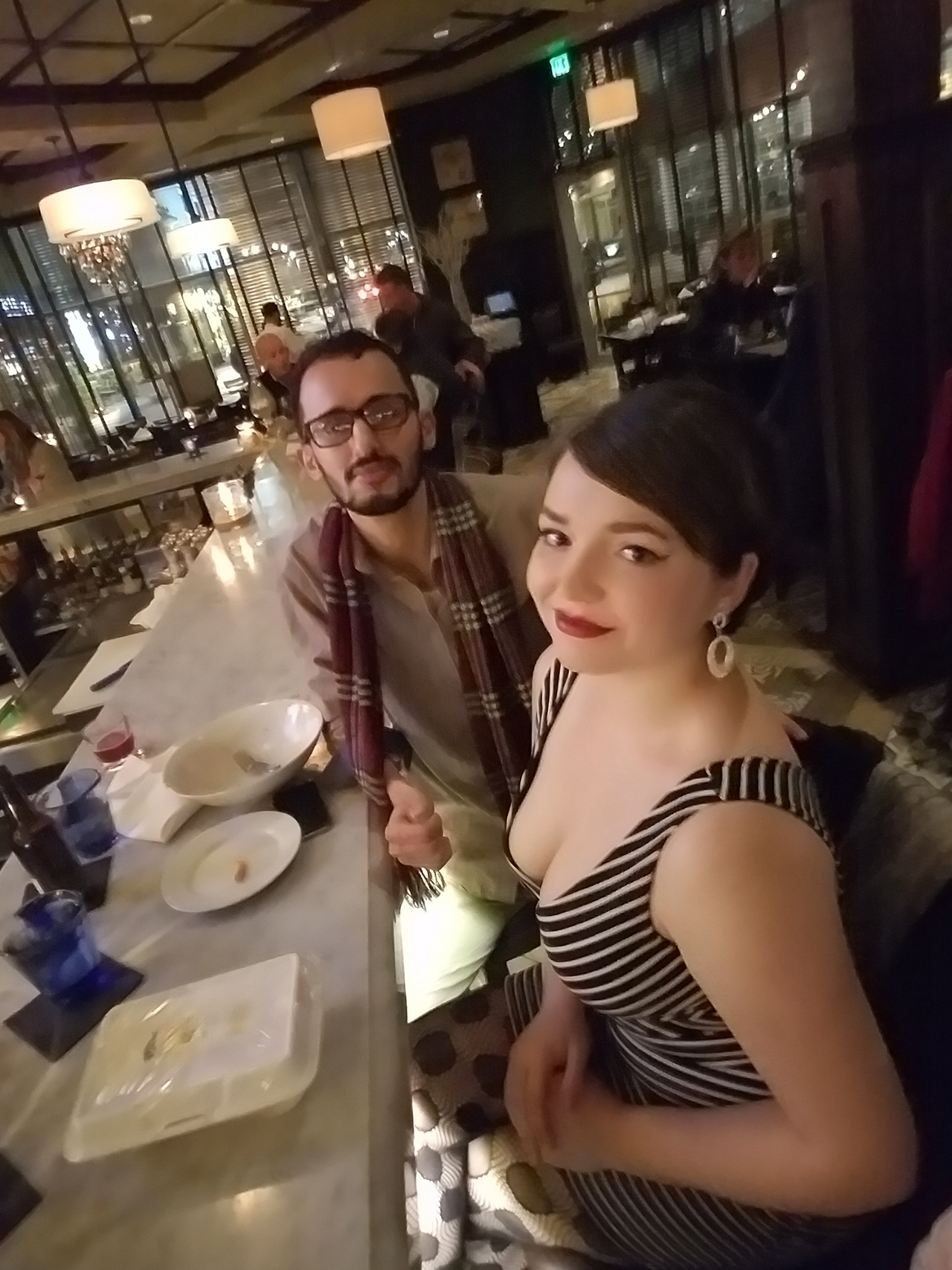a man and woman sitting at a restaurant bar. the woman smiles at the camera, and the man sits at a bit of distance from her with a serious look on his face