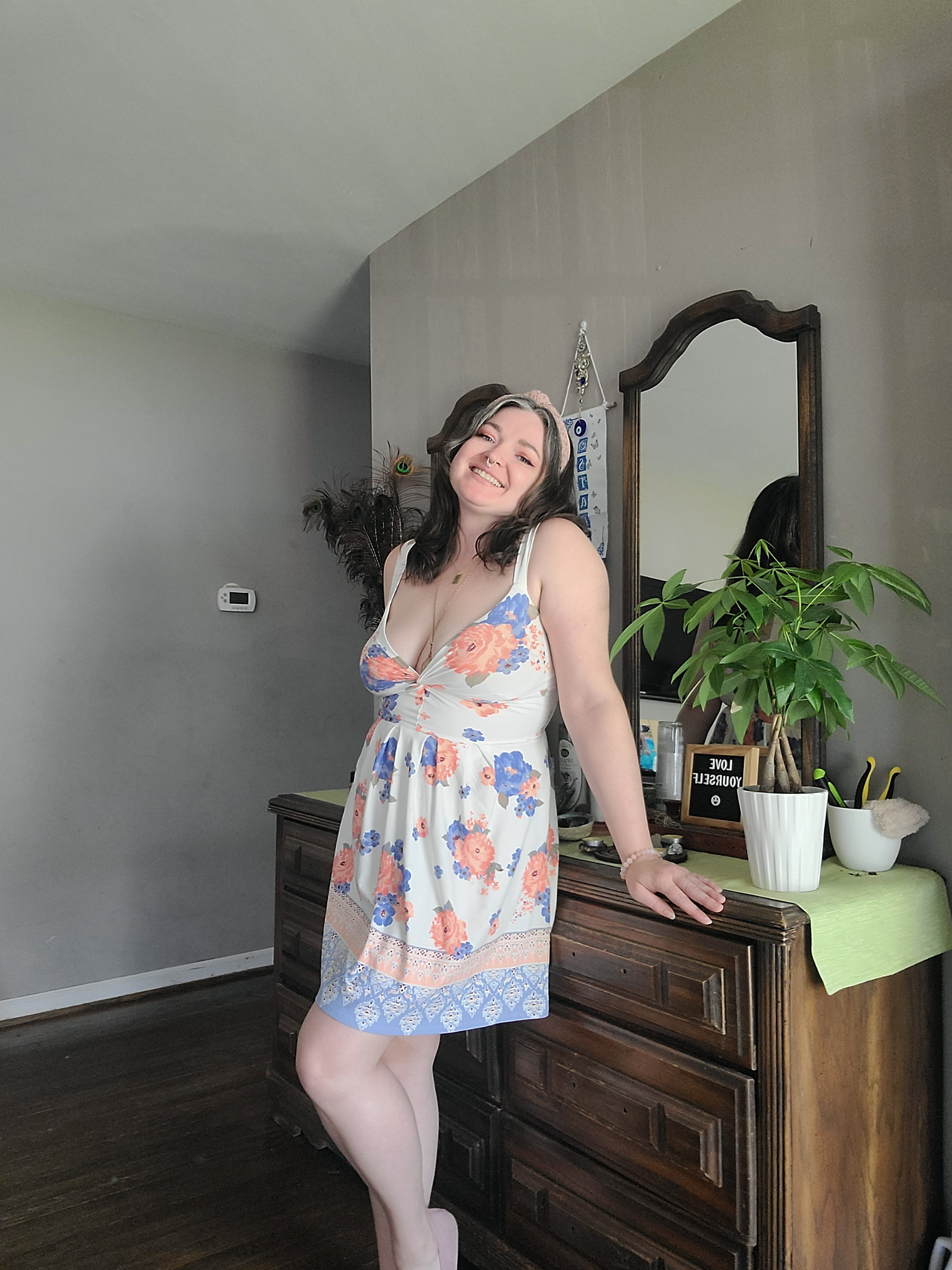 a woman standing in a house in front of a dresser wearing a sundress. she has a radiant smile on her face