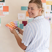 woman facing a colorful wall and holding a color swatch book while smiling over her shoulder