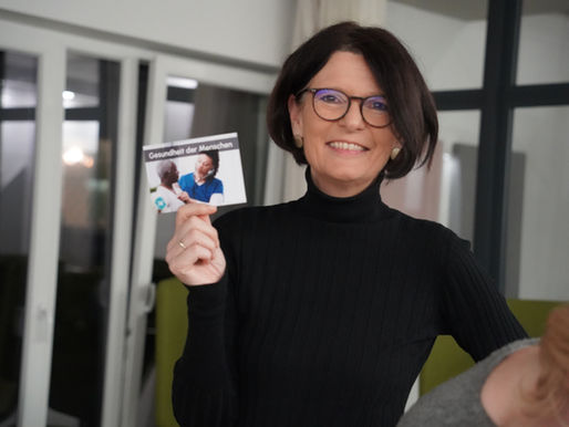 Eine Frau hält eine Karte hoch, die ein Puzzleteil des Serious Games ist hoch. Darauf steht: "Gesundheit der Menschen"