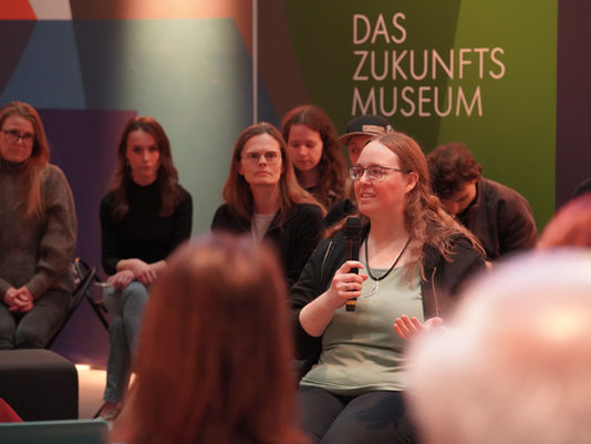 Frau spricht in Mikrofon, umgeben von Zuhörern. Hintergrund mit Schild "Das Zukunfts Museum". Stimmung ist aufmerksam und interessiert.