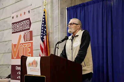 A man named Rick Petro stands at a lecture. A sign saying "Register to Vote" and an American flag are in the background. 