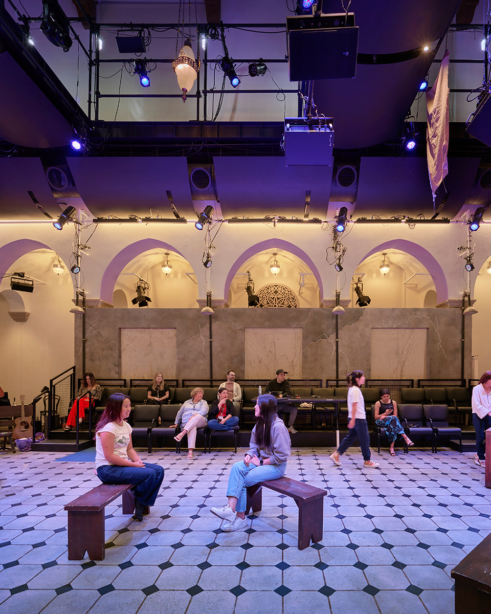 An intimate, flexible studio theatre space within a performing arts venue, featuring stone arches, overhead grid lighting, and students seated on benches.