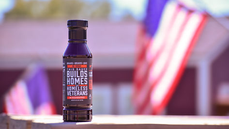 A bottle of Bravo Bravo Que in front of tiny homes and an American flag