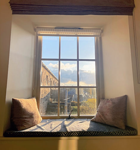 A charming window seat with late afternoon sun streaming through. Two cushions lay either side of the window frame.