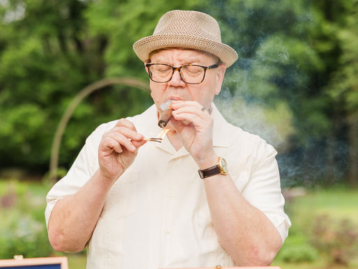 A man wearing a cream shirt and fedora lighting up a cigar.