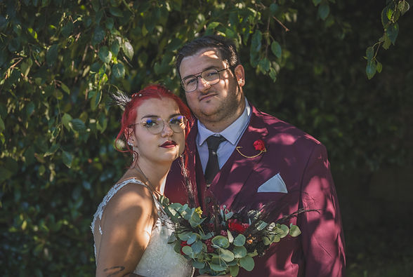 Un couple marié regardant la caméra, bouquet, homme en costume de mariage, arbres en arrière-plan.