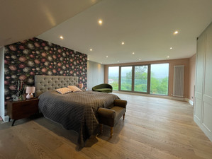 A loft bedroom with a floral accent wall, tufted headboard, large windows, and wooden flooring.