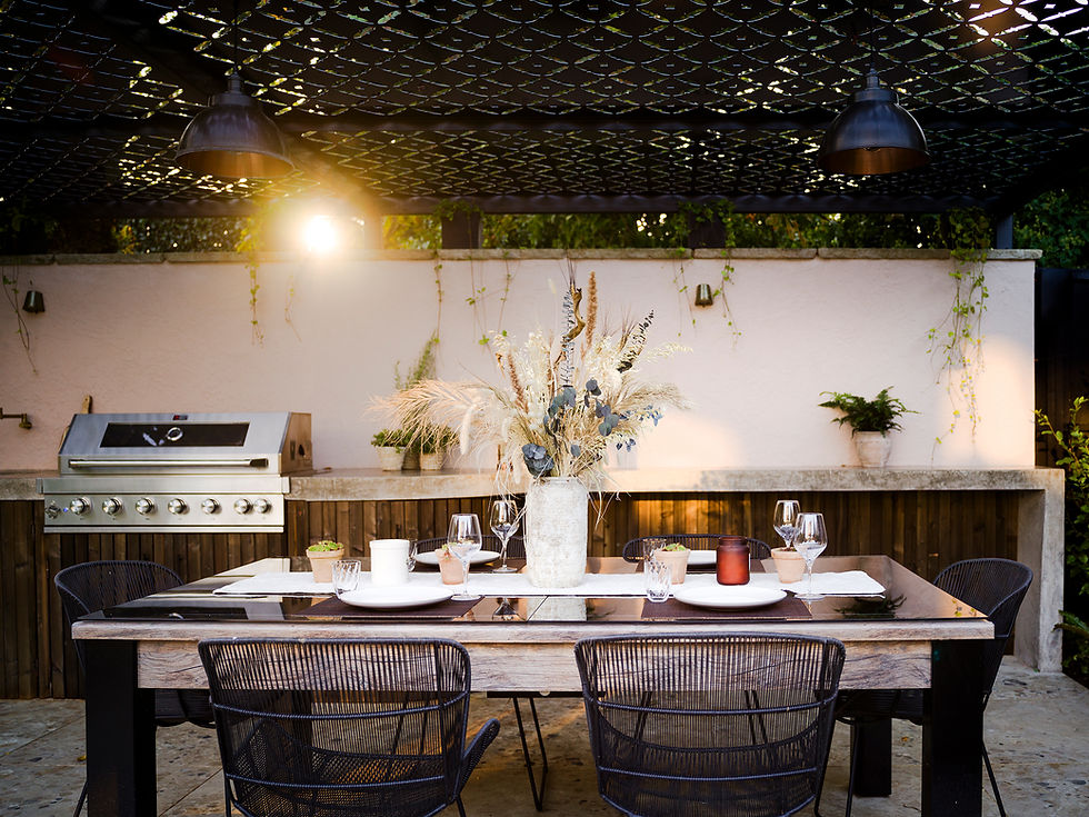 Outdoor dining area with a table set for dinner. There are chairs, a vase of dried flowers, and a grill in the background. Warm lighting.