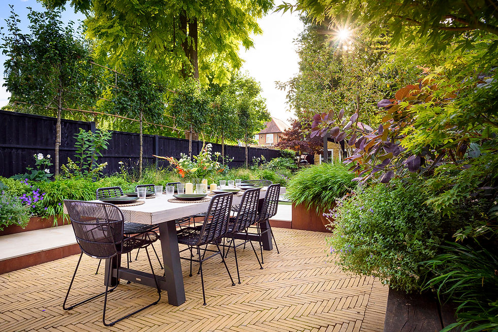 Outdoor dining area with a wooden table set for six, surrounded by lush greenery and flowers. Sunlight filters through trees, creating a serene mood.