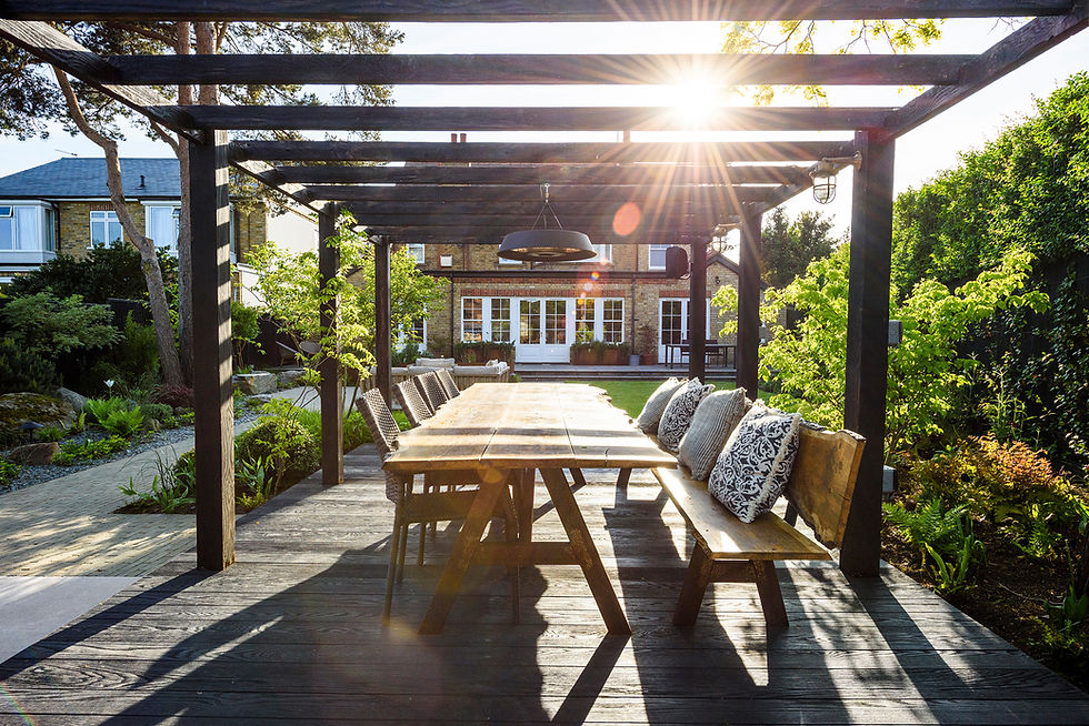 Sunlit garden patio with a long wooden table and cushioned benches under a pergola. Lush greenery surrounds a brick house in the background.