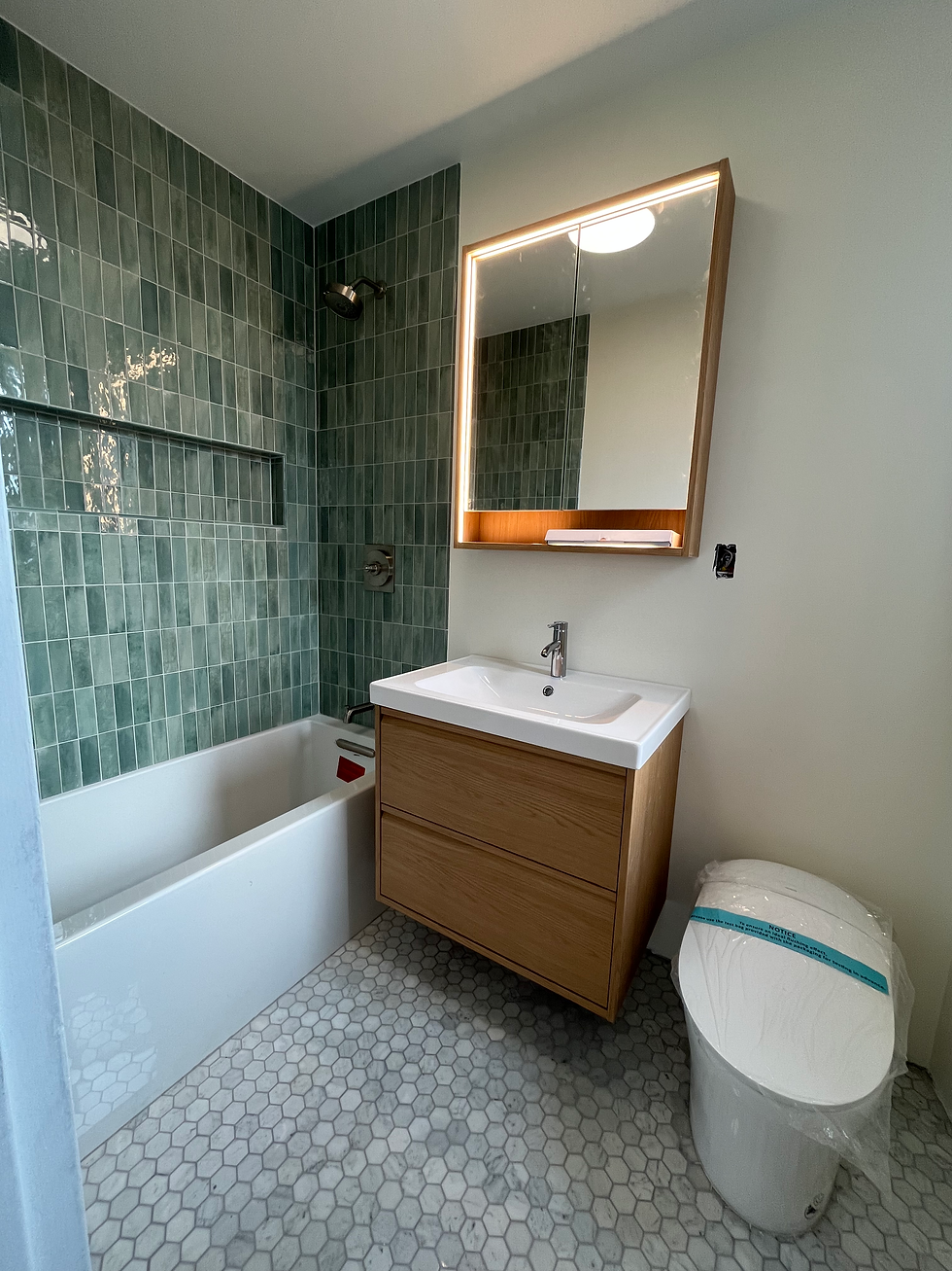 Bathroom Remodeling at Prospect Lefferts Gardens, with green tile wall, white tub, wooden vanity with sink, and toilet. Hexagonal floor tiles and backlit mirror. Calm, modern feel.