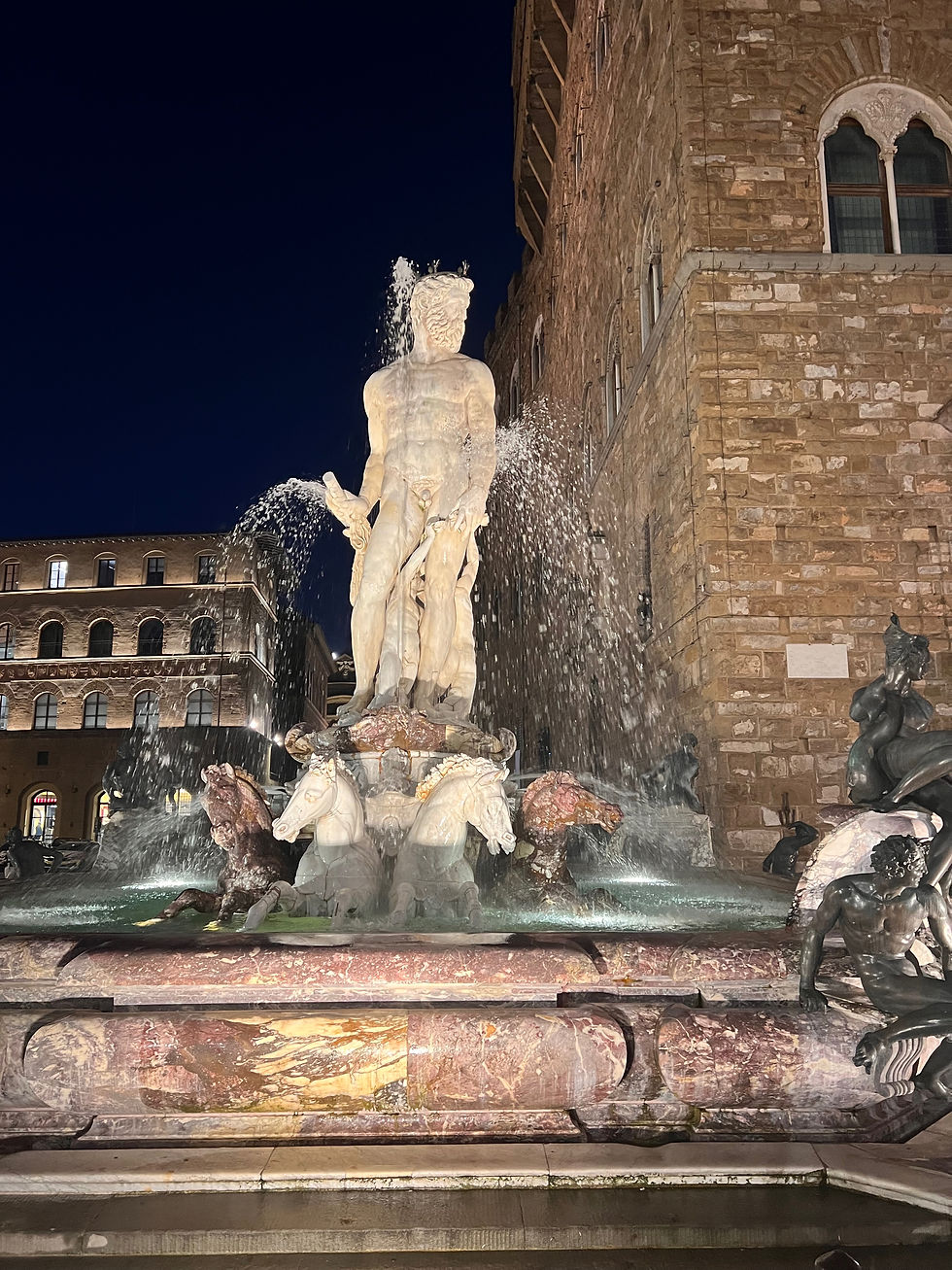 Night fountain with a marble Neptune statue and horse heads spraying water beside a stone building.